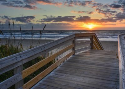 Photo of a boardwalk leading to the beach with the water and sun rising just above the horizon.